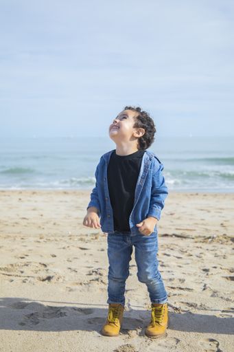 A vertical shot of cheerful little boy in boots playing on the sandy seashore