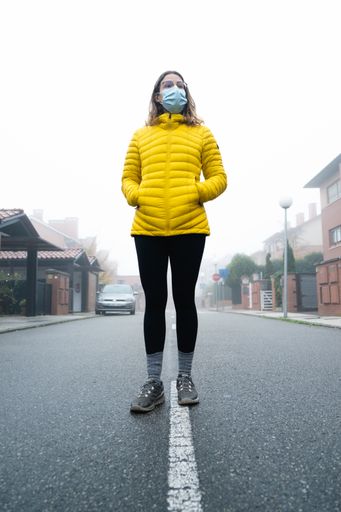 A vertical shot of a young woman standing in the middle of a foggy street wearing a sanitary mask