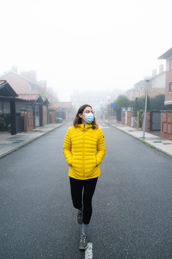 A vertical shot of a young woman walking in the middle of a foggy street wearing a sanitary mask