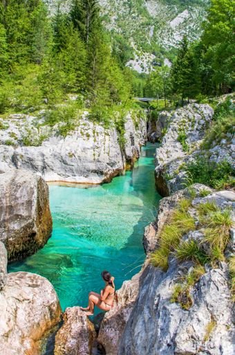 A vertical shot of a female sitting on a rock on the bay of a pure-water river wearing a swimsuit