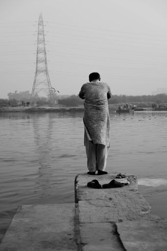 A vertical grayscale of a person standing in front of a water and a power pole with his back