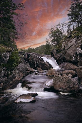 A vertical shot of a waterfall flowing into the rocky river under the cloudy sky