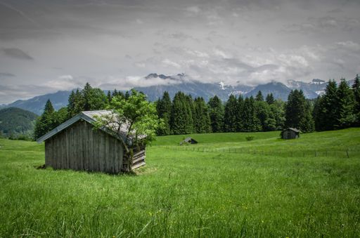 A landscape of a field covered in the grass surrounded by wooden barns and mountains under a cloudy sky