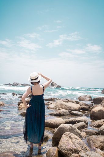 A vertical shot of a female in a blue dress and a straw hat standing in the water and looking