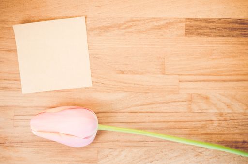 An overhead shot of a pink tulip flower and a blank sticky note on a wooden table with space for your text