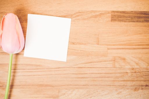 An overhead shot of a pink tulip flower and a blank sticky note on a wooden table with space for your text