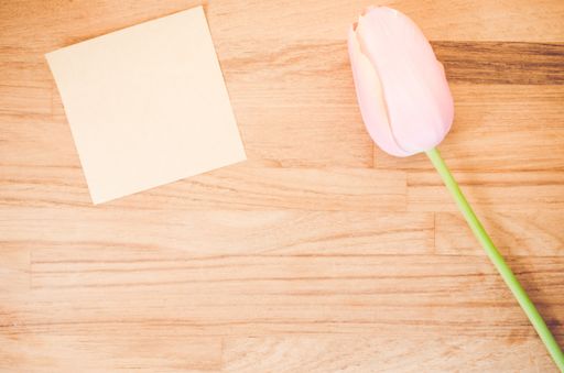 An overhead shot of a pink tulip flower and a blank sticky note on a wooden table with space for your text