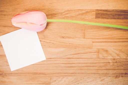 An overhead shot of a pink tulip flower and a blank sticky note on a wooden table with space for your text
