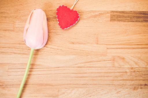 An overhead shot of a pink tulip flower and a handmade felt heart on a wooden table with space for your text