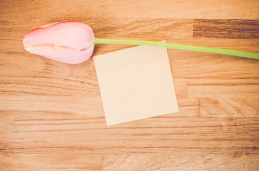 An overhead shot of a pink tulip flower and a blank sticky note on a wooden table with space for your text
