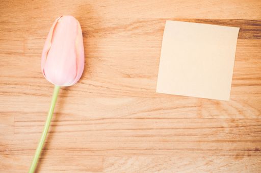 An overhead shot of a pink tulip flower and a blank sticky note on a wooden table with space for your text