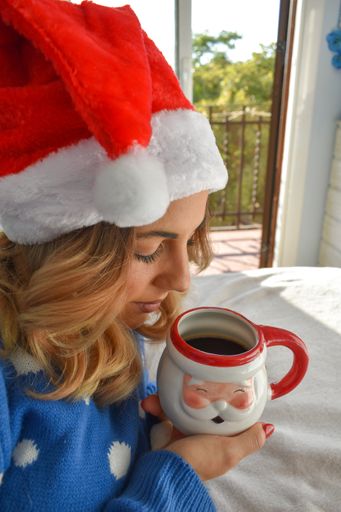 A vertical shot of a Caucasian blonde woman wearing Christmas clothes and drinking coffee from a  mug with Santa Clause