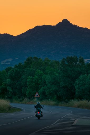 A vertical shot of a biker riding a motorcycle surrounded by hills covered in greenery during the sunset
