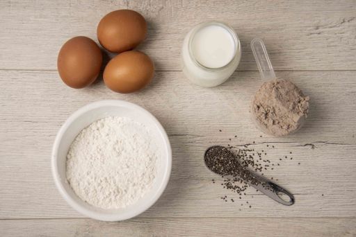 A high angle shot of dough ingredients: flour, eggs, milk, cocoa powder in a scoop, and seeds