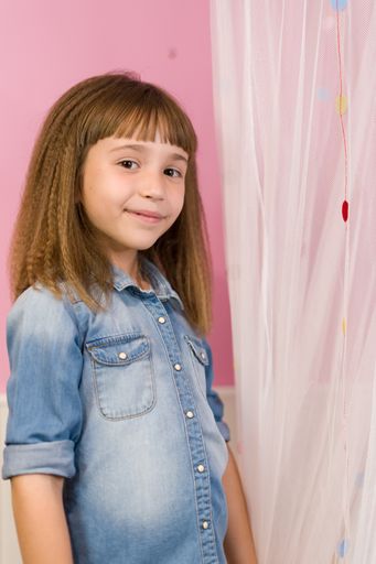 A vertical shot of a cute girl with crimped hair and bangs posing in front of curtains