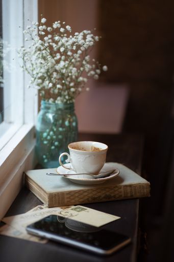 A cup of coffee on a book, a flower vase and a phone on a windowsill