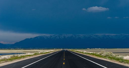 Straight road going towards a very stormy and black sky with the mountains on the horizon under heavy rain.  Angtelope island, Utah, United States of