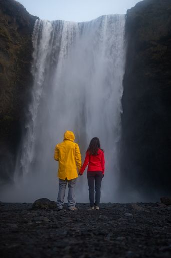 A couple wearing red and yellow clothes standing on a cliff with a waterfall against it