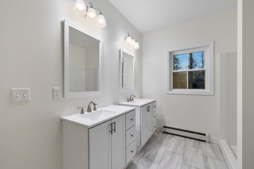 The interior of a white bathroom with wall light fixtures, sinks and cabinets and a baseboard heater under the window
