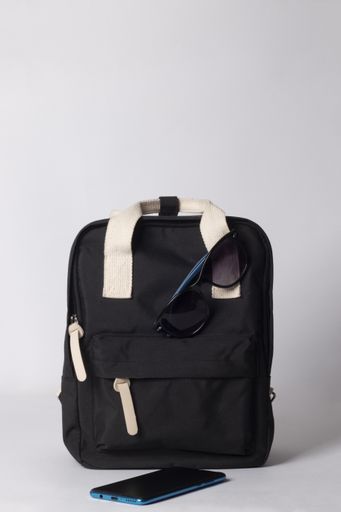A vertical shot of black backpack with sunglasses, and cellphone on white background - travel concept