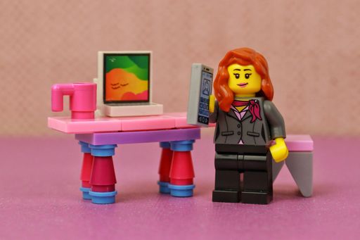 A closeup of a toy red-haired female figure, working from home and holding a phone, standing next to the desk with a computer on it, wearing a suit