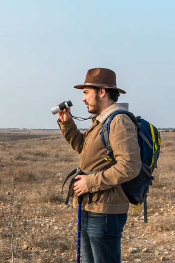 A Caucasian hiker man with a beard, wearing a brown jacket, backpack, looking through binoculars