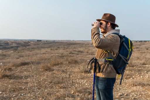 A Caucasian hiker man with a beard, wearing a brown jacket, backpack, looking through binoculars