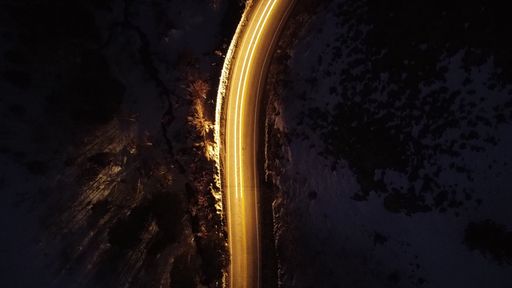 A bird's eye view of a long rural road at night with long exposure yellow car lights illuminating the dark