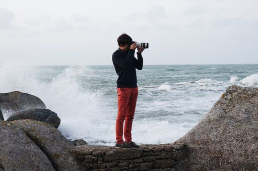 A photographer in red jeans taking a picture while standing on the rocks by the beach.