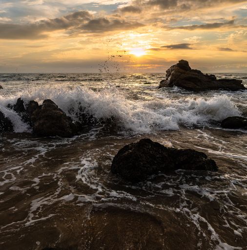 A gorgeous view of vicious sea tides splashing against rocks at the beach under the rising yellow sun