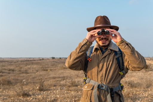 A Caucasian hiker man with a beard, wearing a brown jacket, backpack, looking through binoculars
