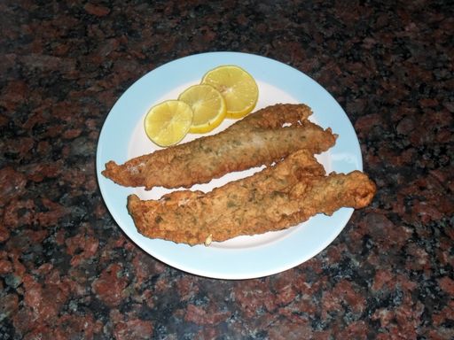 High angle closeup of two fresh fried floured hake fish fillets with lemon slices on a plate with a brown marble background