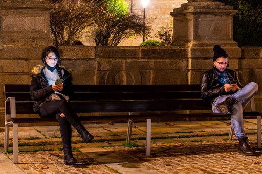 A view of two Caucasian people sitting on a bench while keeping a safe distance with masks