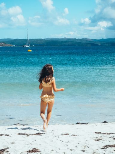A vertical shot of the back of a little girl unrecognizable in a bikini running along the beach towards the water on a sunny day.