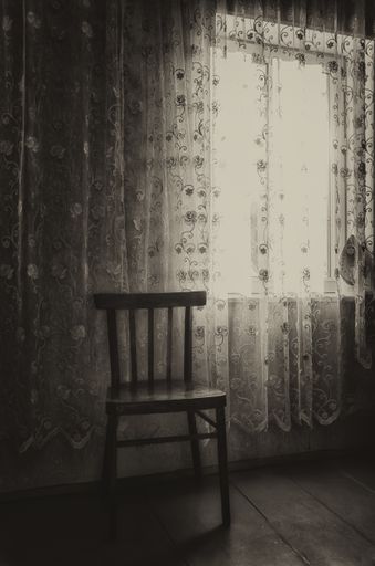 A vertical shot of an empty room with windows, curtains and a wooden chair
