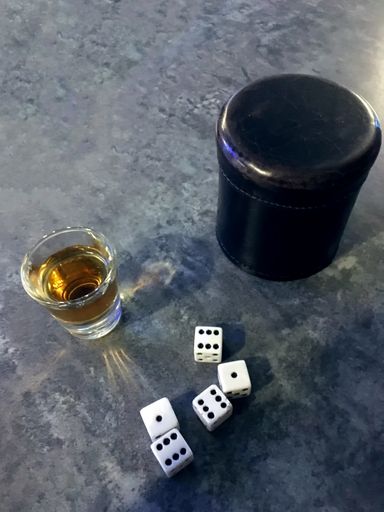 A top shot of dark color liquor with dice and dice cup on a grey mottled bar surface