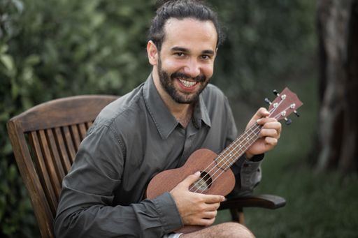 A happy young Hispanic bearded man sitting with a ukulele in a park