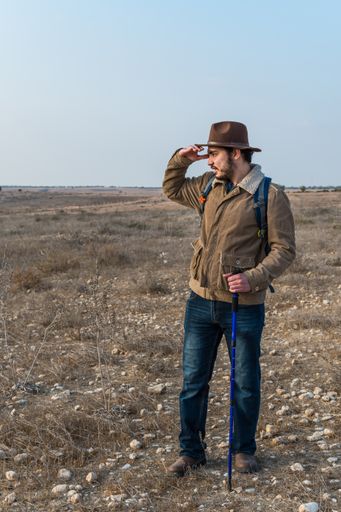 A young hiker with beard wearing brown jacket, backpack and a hipster hat look far away