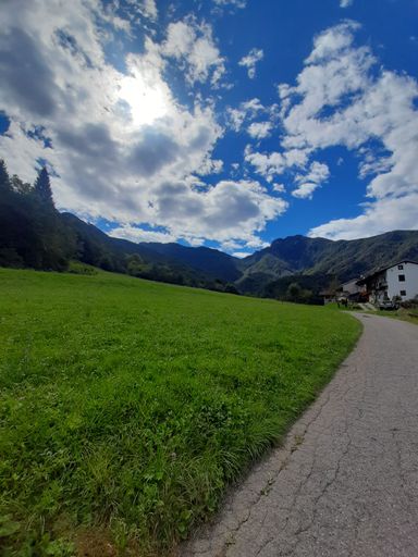 A scenic view of a countryside road against cottages on a green hill under a cloudy blue sky
