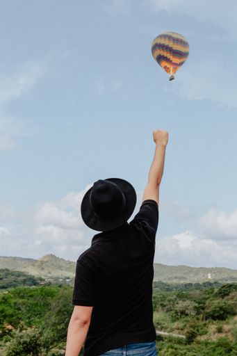 A male standing in the field with his right arm and fist raised into the air and a hot air balloon flying the background