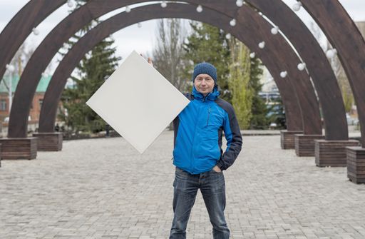 A middle-aged man stands against the background of a cityscape and holds a white blank canvas for painting in his hands