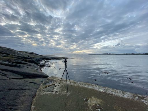 A camera on a stand on the rocky shore surrounded by the sea under a cloudy sky
