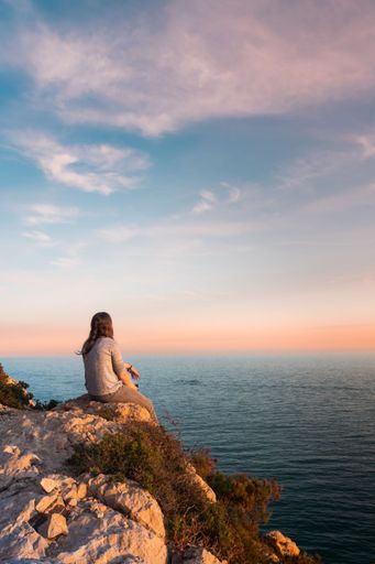 Young woman from behind watching the sea and resting on the edge of a cliff during sunset