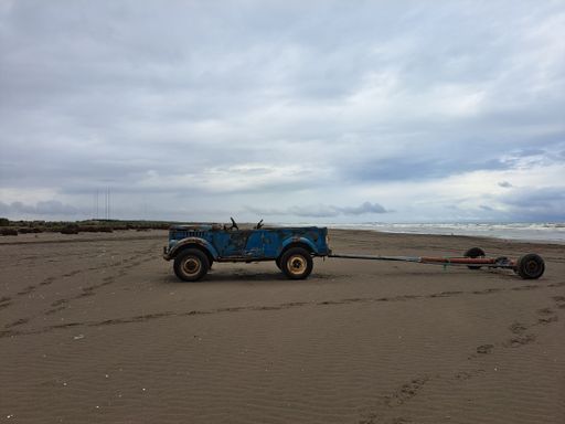 A view of an old blue car with a trailer on sandy beach under cloudy sky