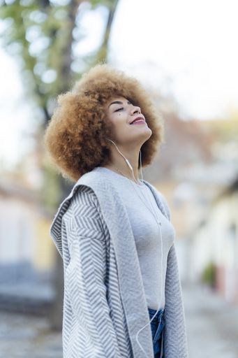 A shallow focus of a young relaxed Hispanic female with curly hair listening to music with headphones in a park