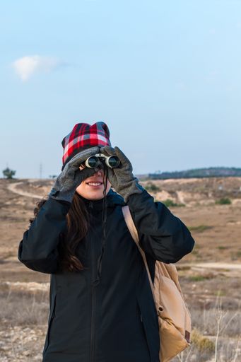 A view of a Caucasian hiker woman wearing a blue jacket, gloves, backpack, looking through binoculars