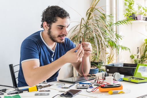 A young repairman of phones and tablets smiles while checking some cables, during his work at home.