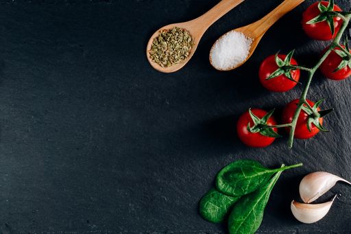An overhead shot of cherry tomatoes, garlic, and wooden spoons full of spice on a dark background