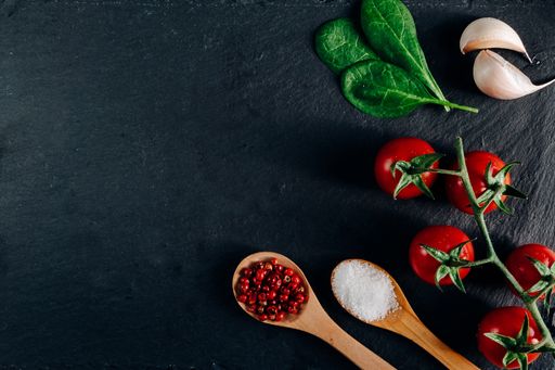 An overhead shot of cherry tomatoes, garlic, and wooden spoons full of spice on a dark background
