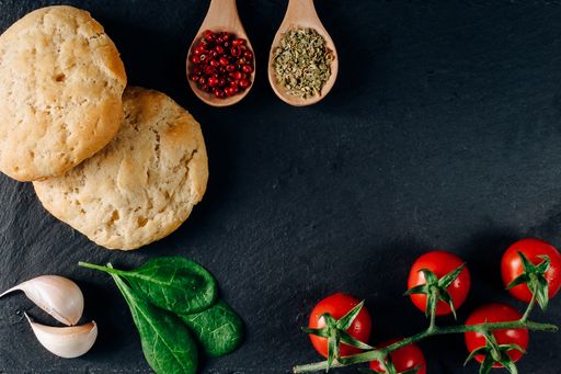 A top view of food ingredients, fresh cherry tomatoes, bread, garlic, spices in tablespoons on a black surface with a copy space